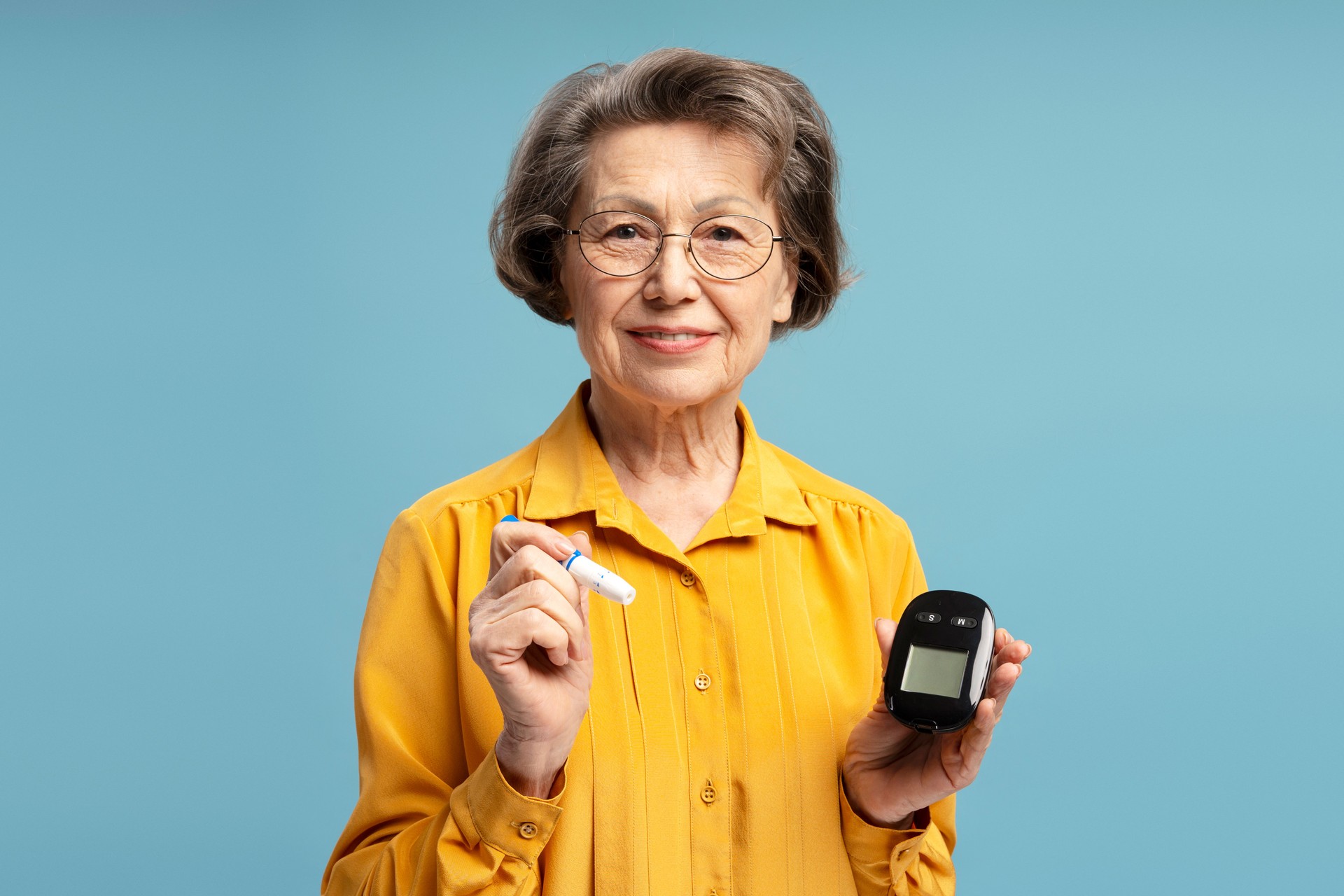 Senior woman holding lancing device and glucometer smiling at camera on blue background